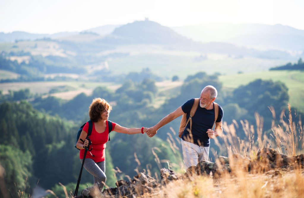 Senior tourist couple with backpacks hiking in nature, holding hands.